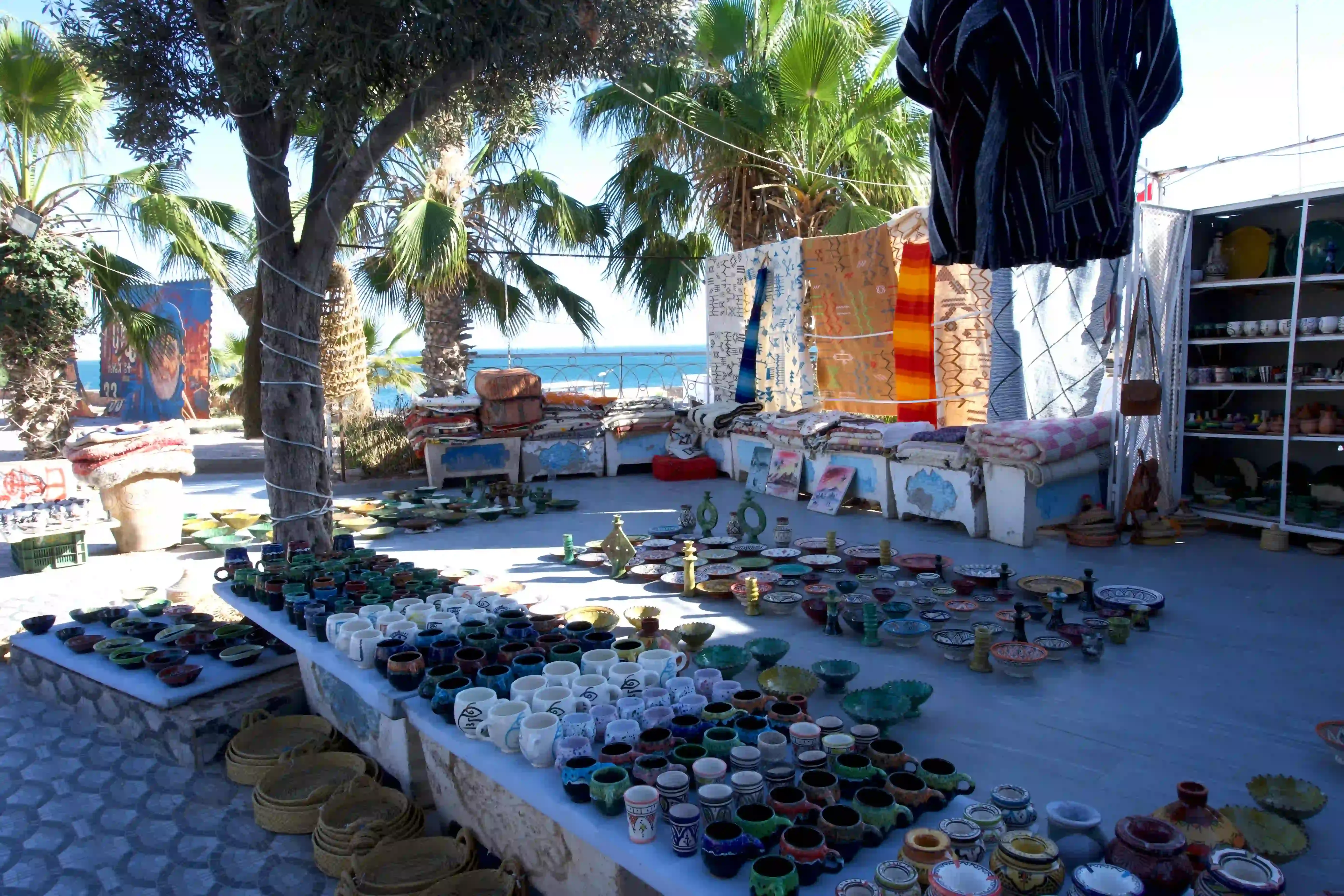 An open-air stall of glazed pottery and tagines beside the sea, with rugs hanging behind and palm trees overhead