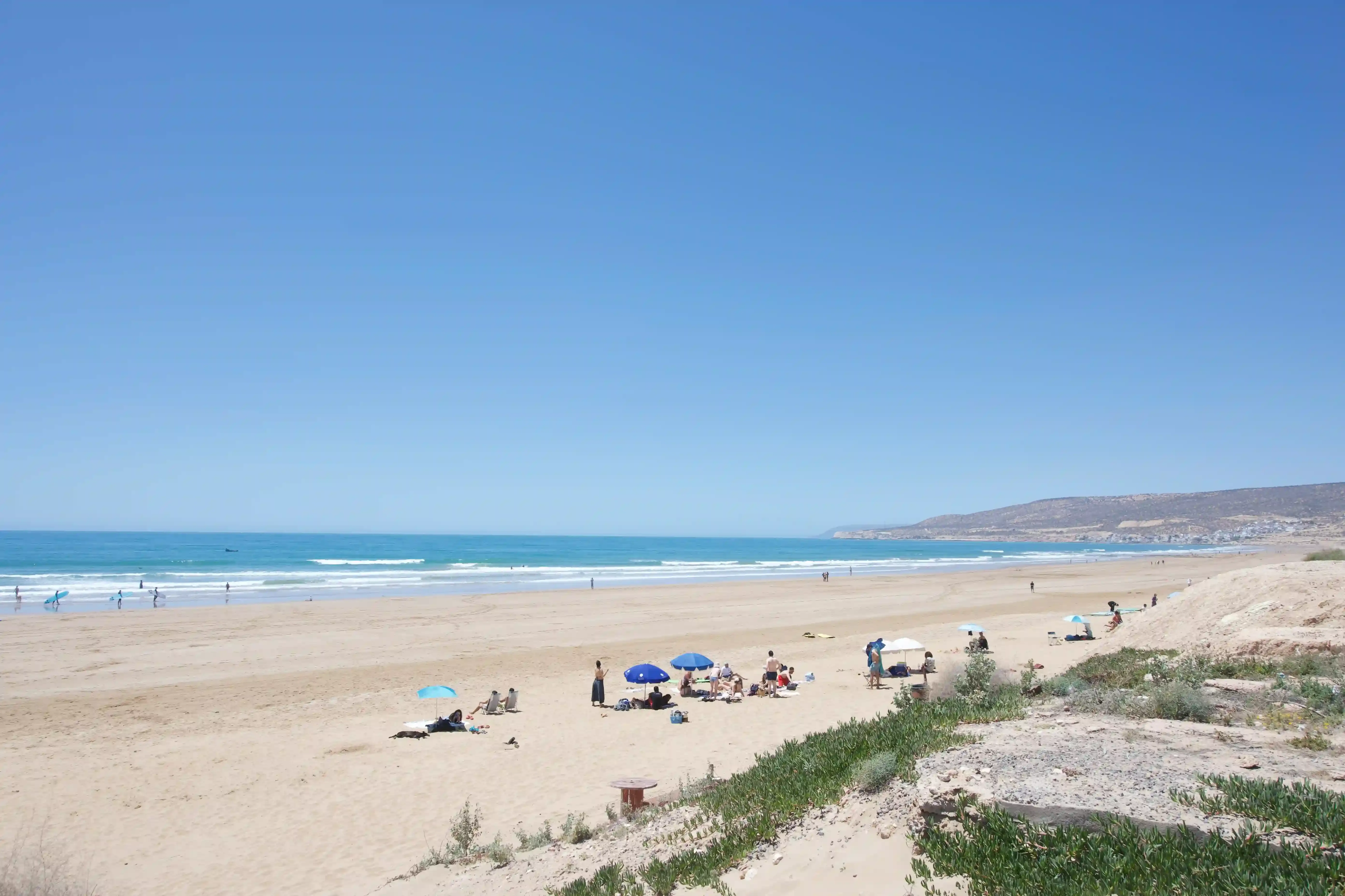 A wide sandy beach under a clear sky, a few parasols near the shoreline and hills at the far end of the bay